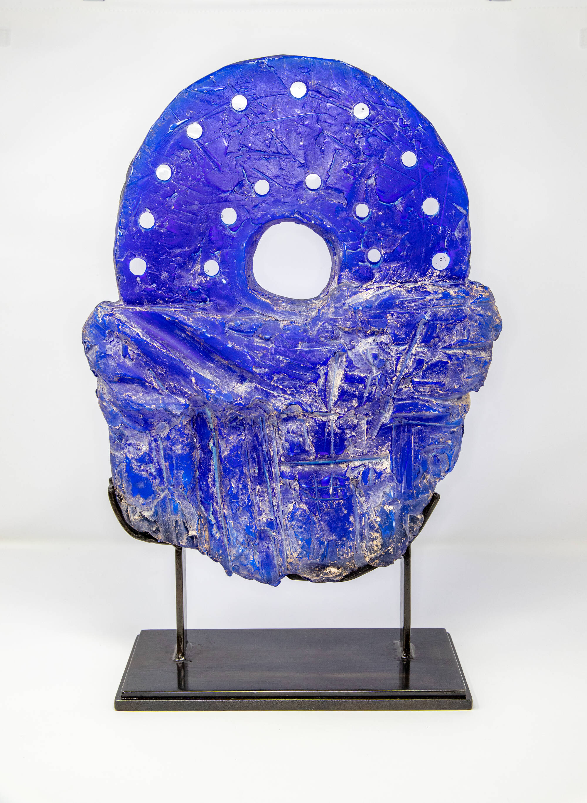 Blue glass piece that sits in metal stand. Glass is cut in a circular, or wheel, shape at the top and left more raw at the bottom.
